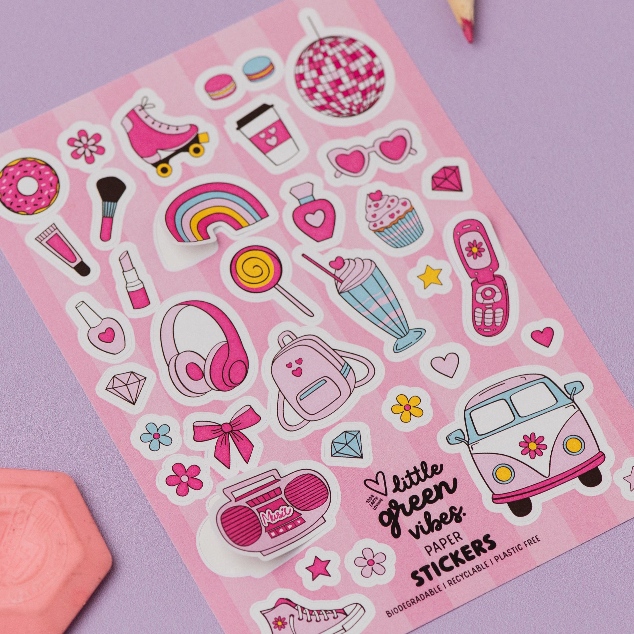 Girly Dreamworld Pink Sticker Sheet featuring cute pink and white illustrations including roller skates, disco ball, headphones, rainbow, hearts, milkshake, cupcake, vintage van, and other girly icons on a pink background, displayed on purple surface with colored pencils and pink geometric shape