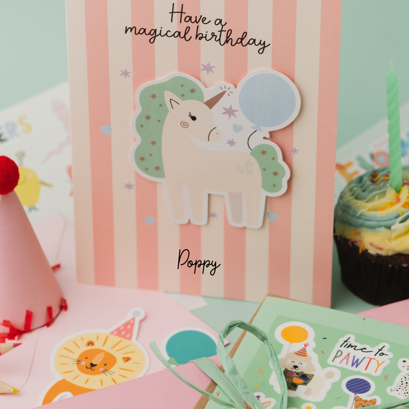 Personalised Magical Birthday Unicorn Sticker & Colouring Kids Birthday Card featuring a white unicorn with mint green mane holding a blue balloon against pink and white striped background. Card displays 'Have a magical birthday' text with scattered silver stars and personalized name.