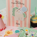 Personalised Magical Birthday Unicorn Sticker & Colouring Kids Birthday Card featuring a white unicorn with mint green mane holding a blue balloon against pink and white striped background. Card displays 'Have a magical birthday' text with scattered silver stars and personalized name.