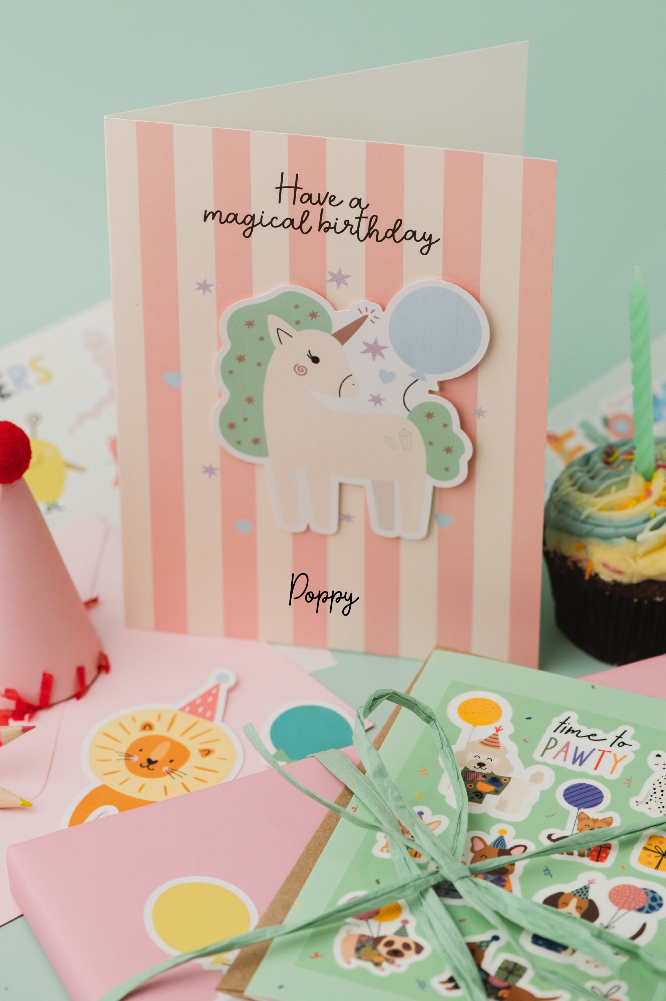 Personalised Magical Birthday Unicorn Sticker & Colouring Kids Birthday Card featuring a white unicorn with mint green mane holding a blue balloon against pink and white striped background. Card displays 'Have a magical birthday' text with scattered silver stars and personalized name.
