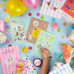 Circus Multi Sticker Kids & Colouring Birthday Card displayed with colorful balloons, confetti dots, and party supplies on a light blue background. Multiple greeting cards featuring circus-themed stickers and coloring designs spread across the surface with festive decorations.
