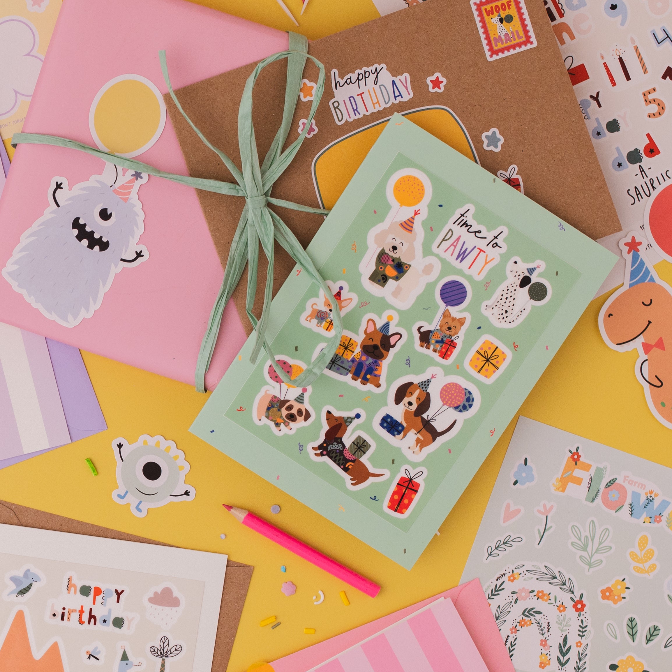 Dog Multi Sticker & Colouring Kids Birthday Card featuring playful stickers with cartoon dogs, colorful birthday decorations, and activity sheets spread on yellow background. Card set includes mint green sticker sheet, pink and kraft paper envelopes, and party-themed decorative elements.