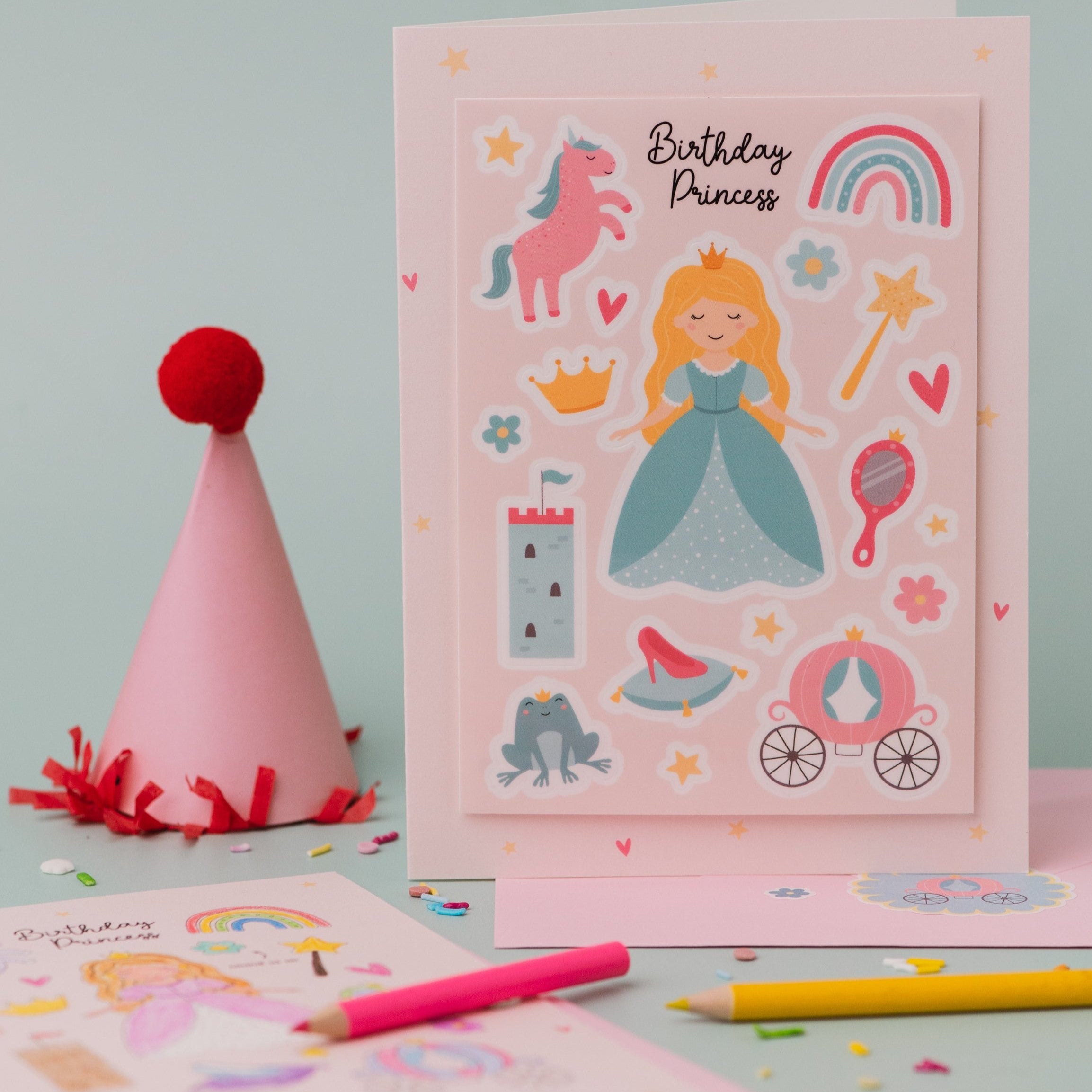 Princess Sticker & Colouring activity Birthday Card featuring a pink design with magical princess illustrations including a unicorn, rainbow, castle, crown, carriage, and fairy tale elements, displayed alongside colourful pencils and a pink party hat