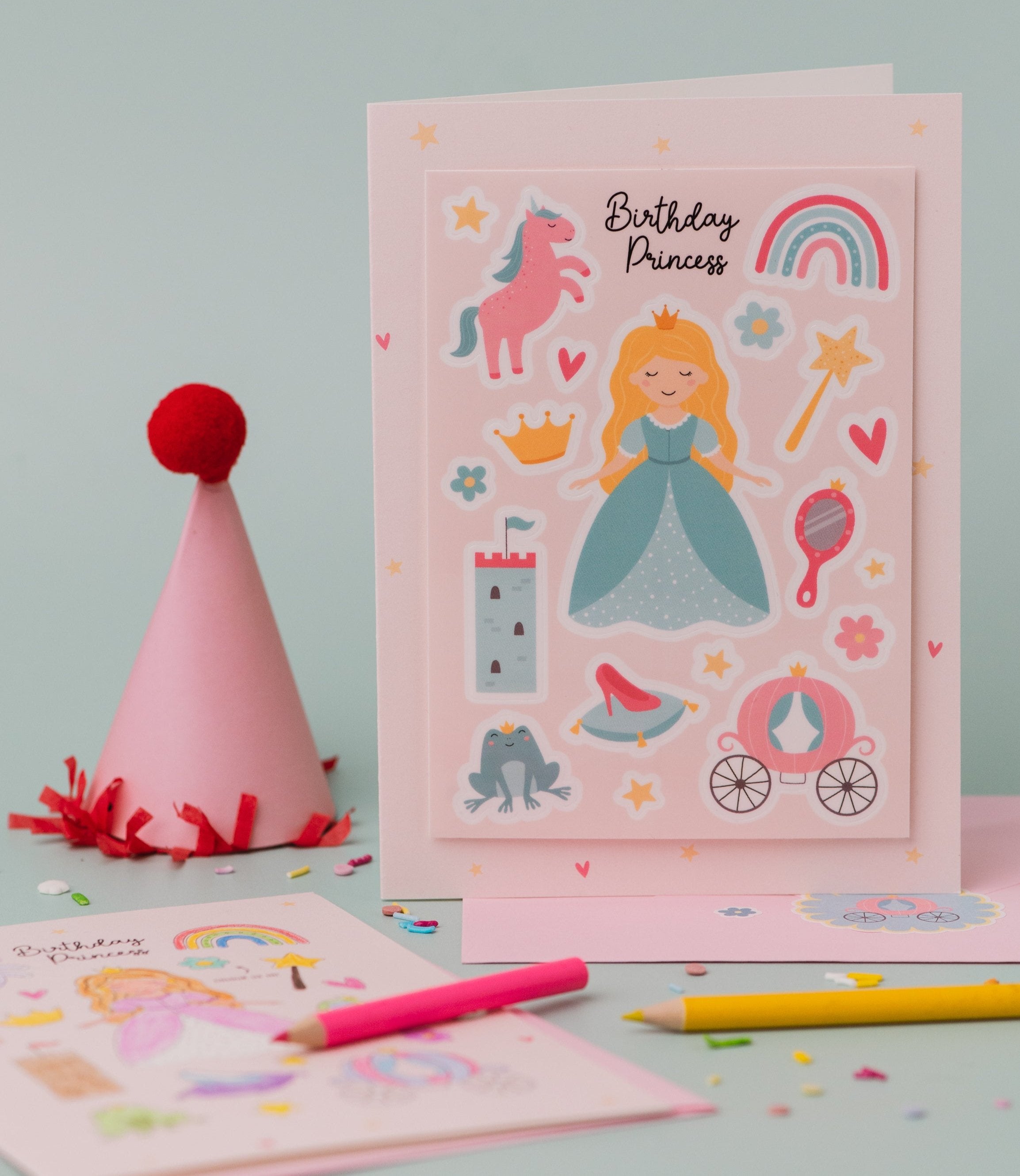 Princess Sticker & Colouring activity Birthday Card featuring a pink design with magical princess illustrations including a unicorn, rainbow, castle, crown, carriage, and fairy tale elements, displayed alongside colourful pencils and a pink party hat