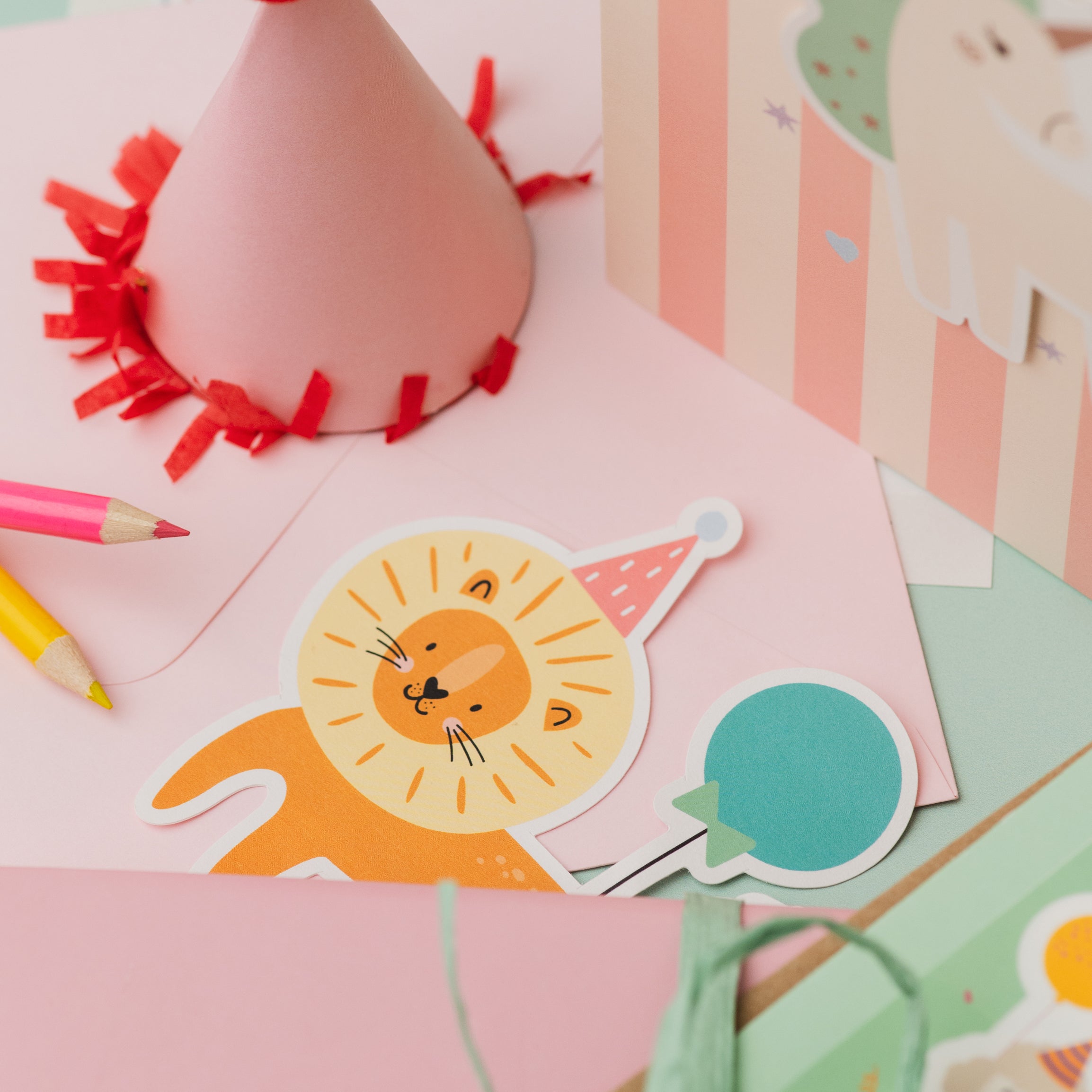 Lion Personalised Sticker & Colouring Kids Birthday Card featuring a cute cartoon lion sticker with party hat on pink background, alongside colorful pencils and a festive red-trimmed party hat decoration. Pastel striped background with playful birthday party elements.