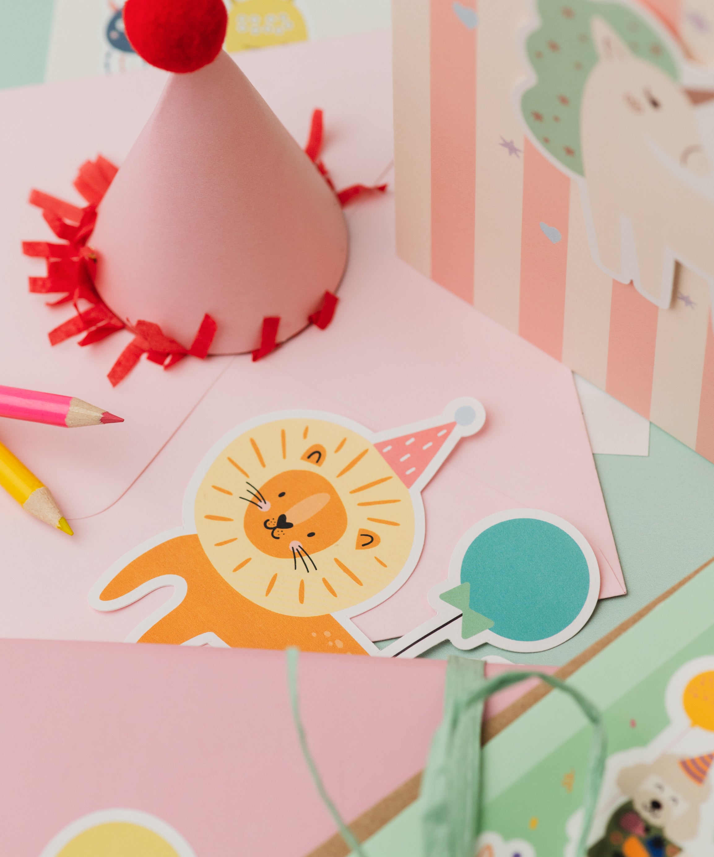 Lion Personalised Sticker & Colouring Kids Birthday Card featuring a cute cartoon lion sticker with party hat on pink background, alongside colorful pencils and a festive red-trimmed party hat decoration. Pastel striped background with playful birthday party elements.