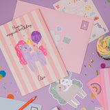 Personalised Magical Birthday Unicorn Sticker & Colouring Kids Birthday Card featuring a hand-drawn purple unicorn with pink mane holding a balloon on pink striped background, displayed with matching envelopes, stickers, and celebratory cupcake on purple surface with scattered confetti and rainbow decorations