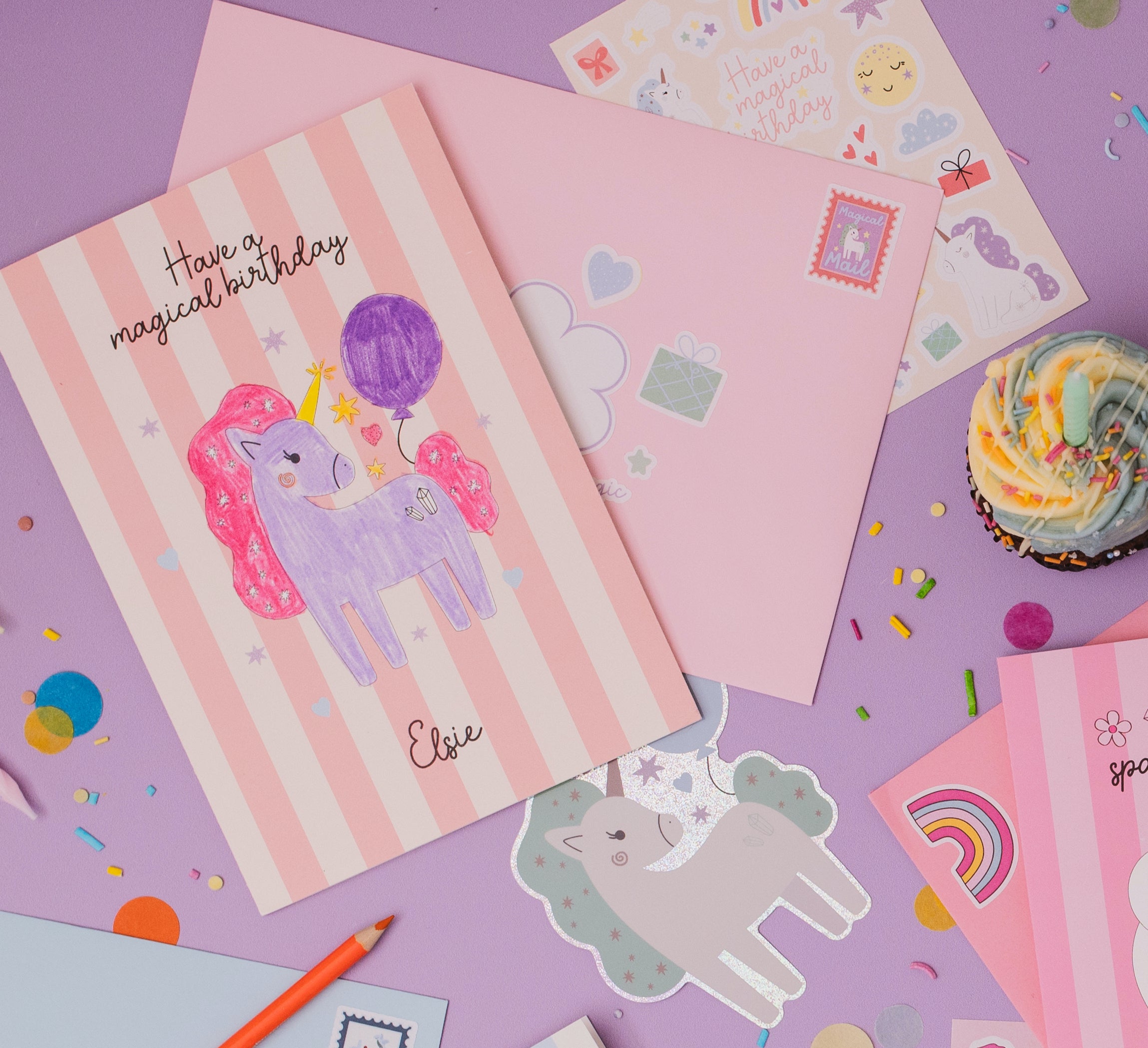 Personalised Magical Birthday Unicorn Sticker & Colouring Kids Birthday Card featuring a hand-drawn purple unicorn with pink mane holding a balloon on pink striped background, displayed with matching envelopes, stickers, and celebratory cupcake on purple surface with scattered confetti and rainbow decorations