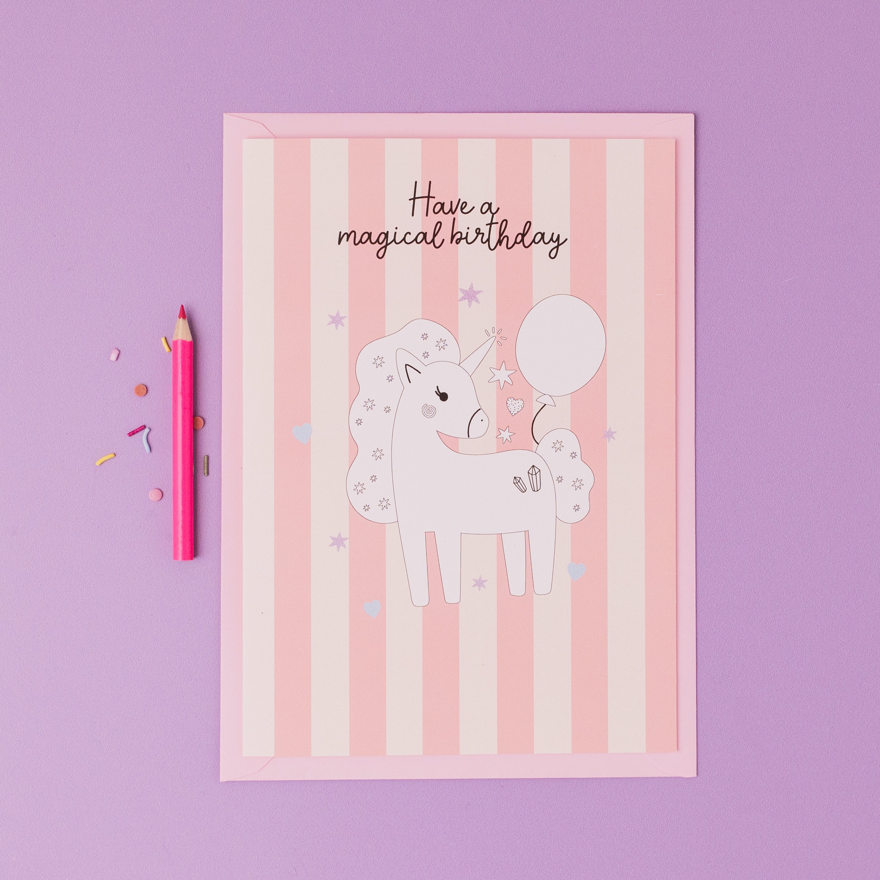 Personalised Magical Birthday Unicorn Sticker & Colouring Kids Birthday Card featuring a white unicorn illustration holding a balloon on pink striped background with silver stars, accompanied by a pink pencil and confetti on purple surface