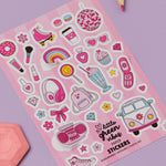 Girly Dreamworld Pink Sticker Sheet featuring cute pink and white illustrations including roller skates, disco ball, headphones, rainbow, hearts, milkshake, cupcake, vintage van, and other girly icons on a pink background, displayed on purple surface with colored pencils and pink geometric shape