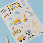 Construction Sticker Sheets featuring colorful construction vehicles and safety signs on white paper, including yellow bulldozers, cranes, dump trucks, and warning barriers, displayed on a light blue background with a green hexagonal object and pencils nearby