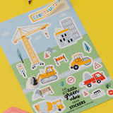 Construction Sticker Sheets featuring colorful construction vehicles and equipment on a light blue and green background, including crane, bulldozer, cement mixer, and traffic signs. Two wooden pencils visible against yellow background. Eco-friendly paper stickers arranged in a playful scene with buildings and roads.