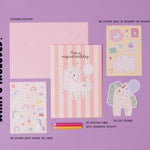 Personalised Magical Birthday Unicorn Sticker & Colouring Kids Birthday Card displayed with matching sticker sheets featuring cute unicorns, rainbows, and stars on pink striped background. Set includes coloring pencils and activity sheets arranged on purple surface.